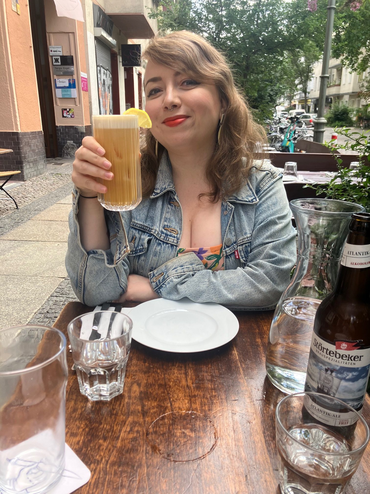 A picture of me at a table, with a glass and a plate in front of me. I have long, wavy brown hair, white skin, and I'm wearing full make-up including orange lipstick and winged eyeliner. I'm wearing a denim jacket and I'm holding a dusky orange cocktail in my right hand. My other arm is folded in front of me. I'm smiling at the camera