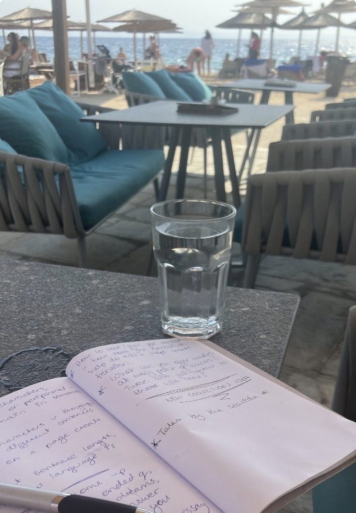 A cafe by the seaside in Greece. In the background are multiple parasols, under which stand multiple people, just out of focus. In the mid ground are chairs with menus atop them, next to which are benches covered in turquoise cushions. In the foreground is a grey table in focus, on top of which are a glass of water and an open notebook, with a pen lying across one corner, 