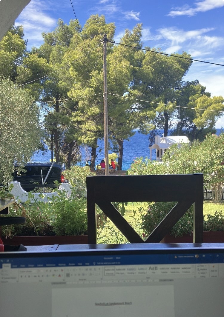 A shot of a Greek garden, with a large tree taking up most of the shot, set against a cloudy, blue sky and a sapphire-coloured sea. In the foreground is a chair, and the top of a laptop, with the beginning of a new story on it.