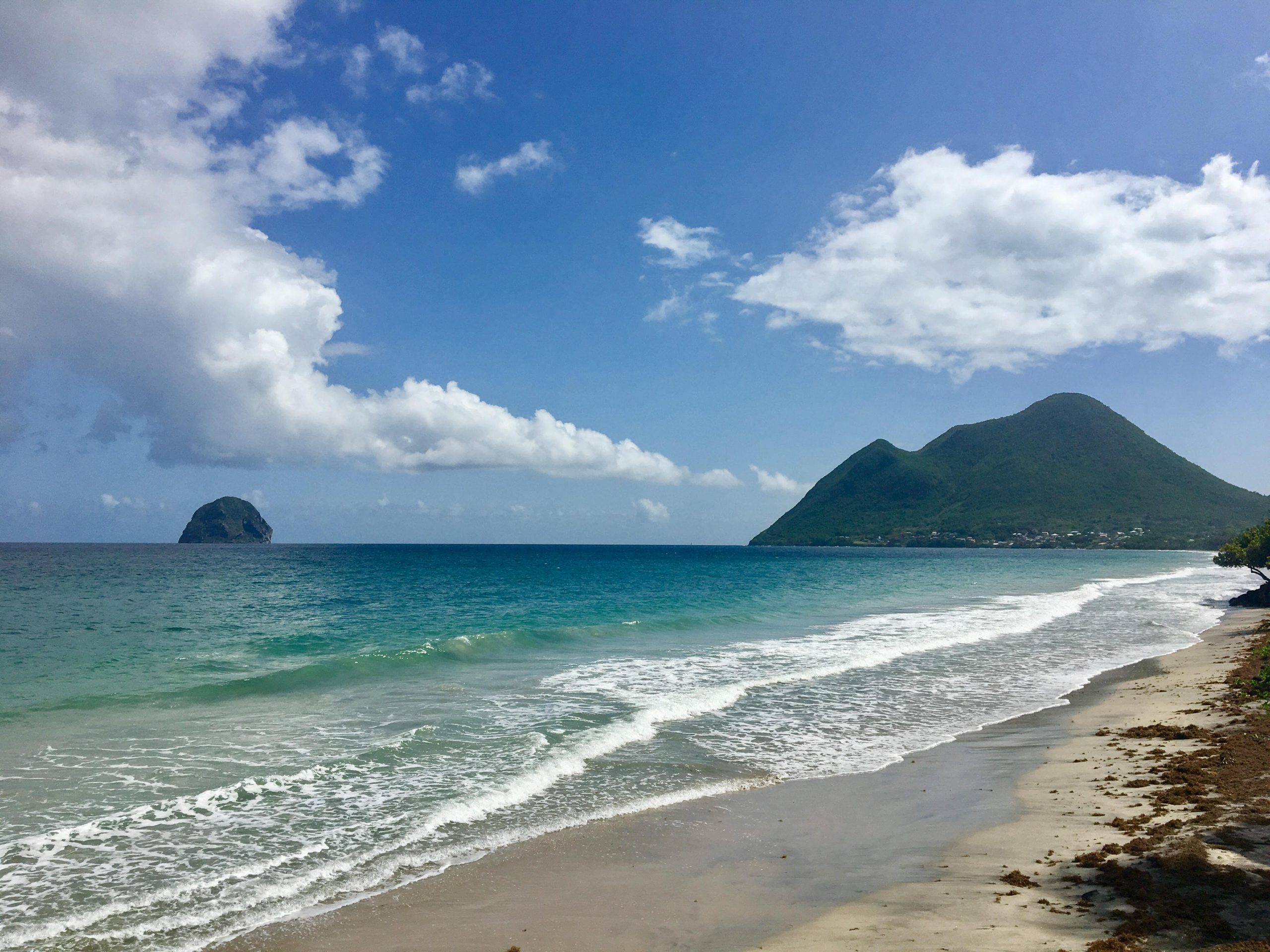 A shot of the seaside. In the bottom right corner the beach is visible. with piles of seaweed along it. The sea is rolling in from the left of the picture, turquise blue under a lightly cloudy blue sky. A small rock is visible in the distance on the left of the picture, as well as a larger hill towards the right-hand side, with multiple peaks.